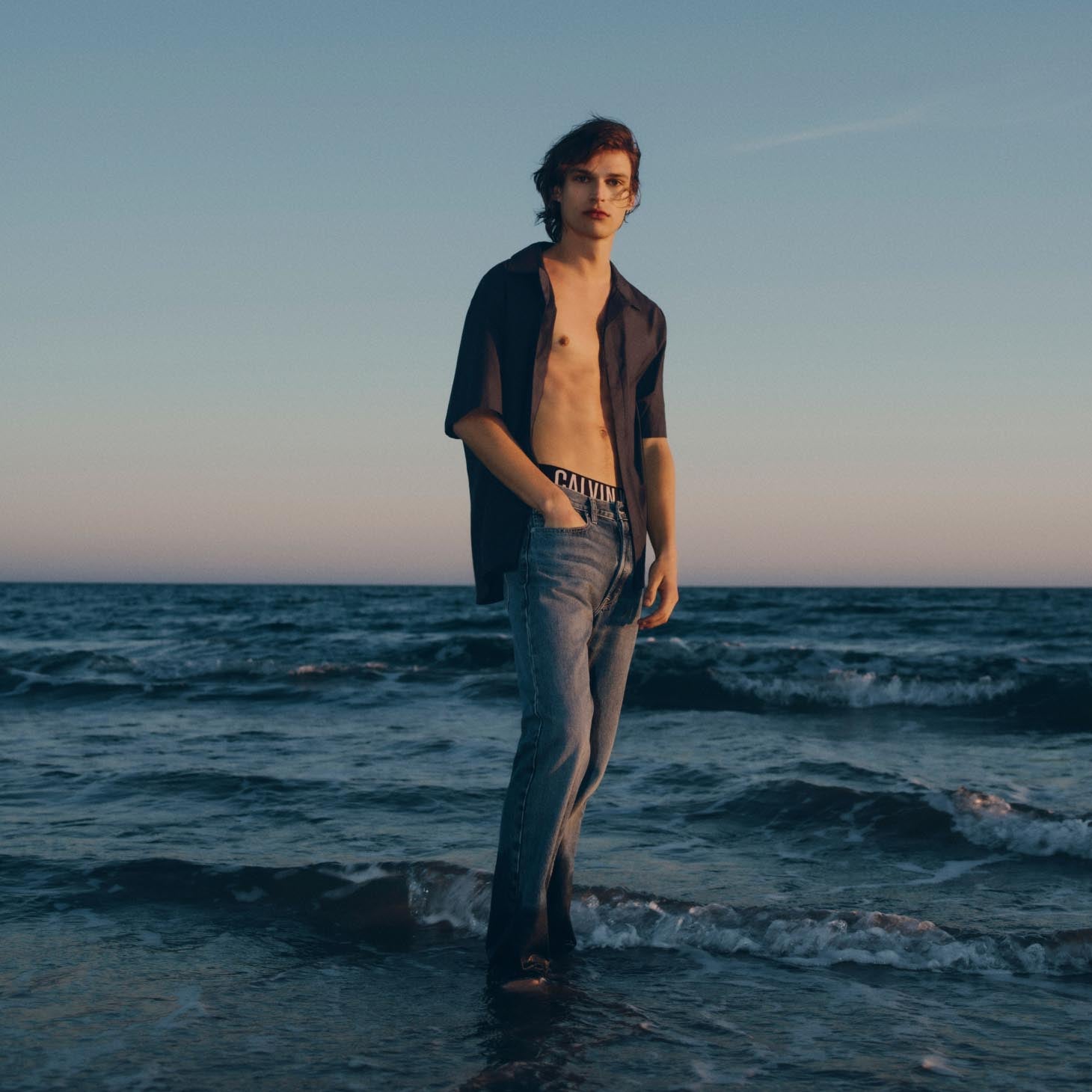 Man standing in the ocean wearing an unbutton short sleeve shirt and denim jeans