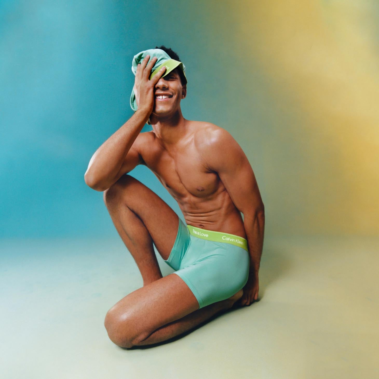 Man kneeling on the ground wearing light green underwear trunks and holding a pair of underwear over his eye