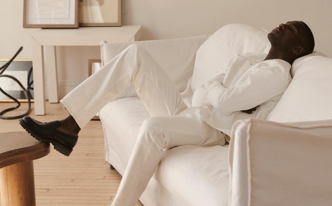 Man sitting on a couch with his leg up on a table wearing a white blazer and white jeans