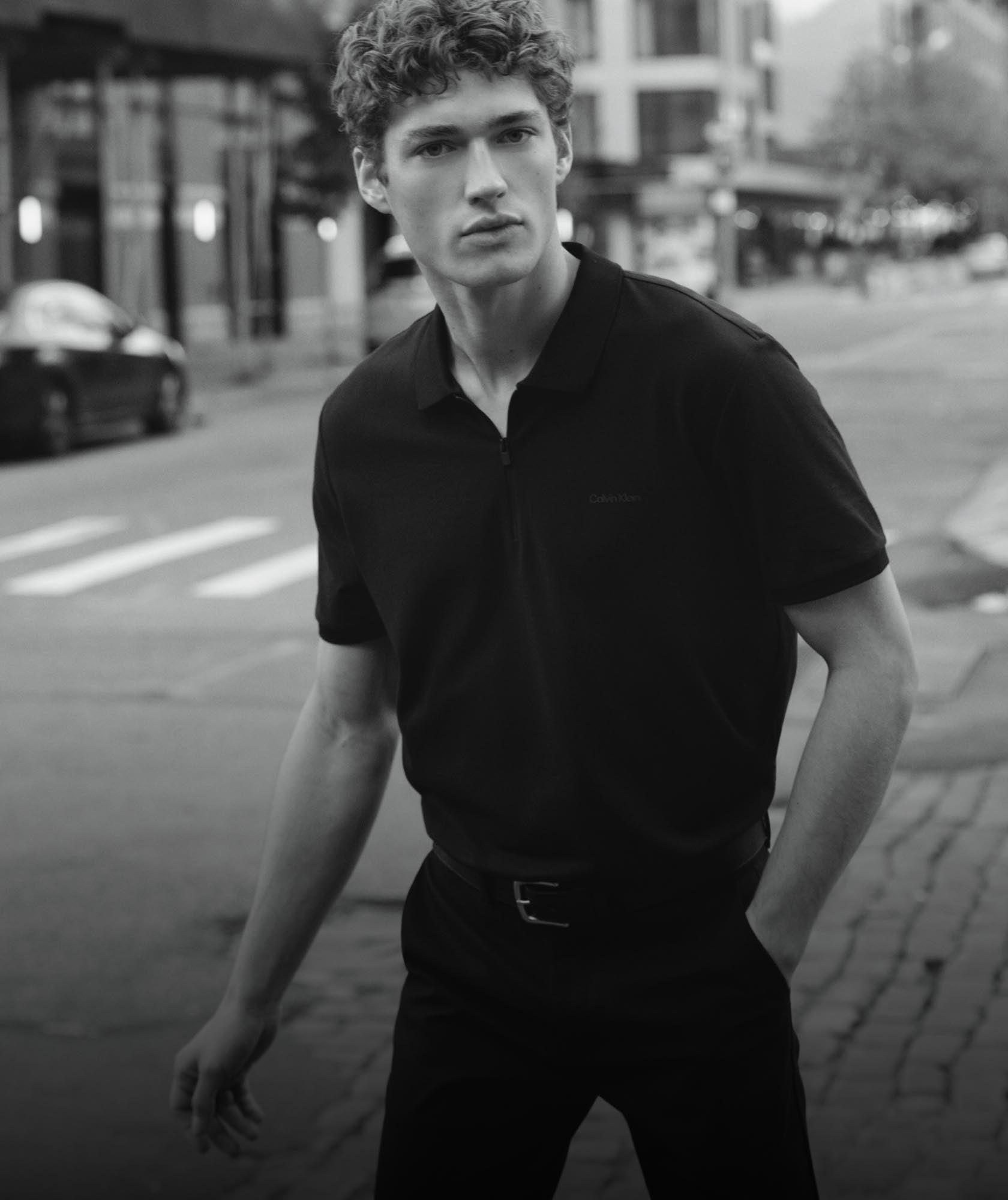 Man on a city street with his hand in his pocket wearing a black zip polo