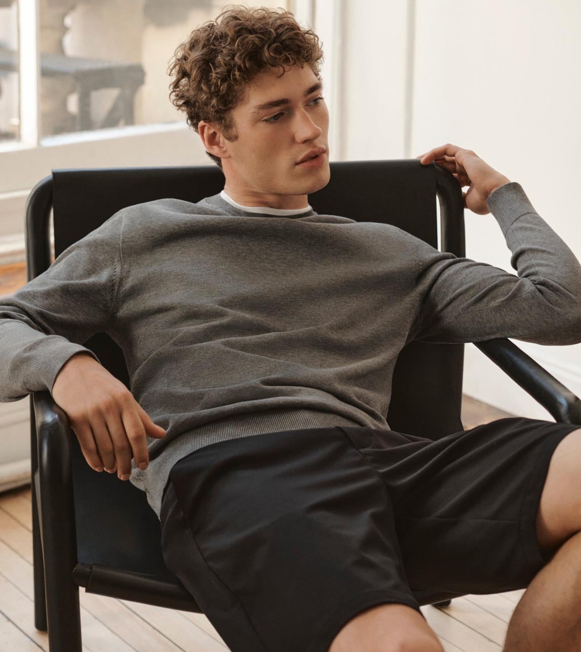 Man leaning back on a chair wearing a grey sweater and black tailored shorts