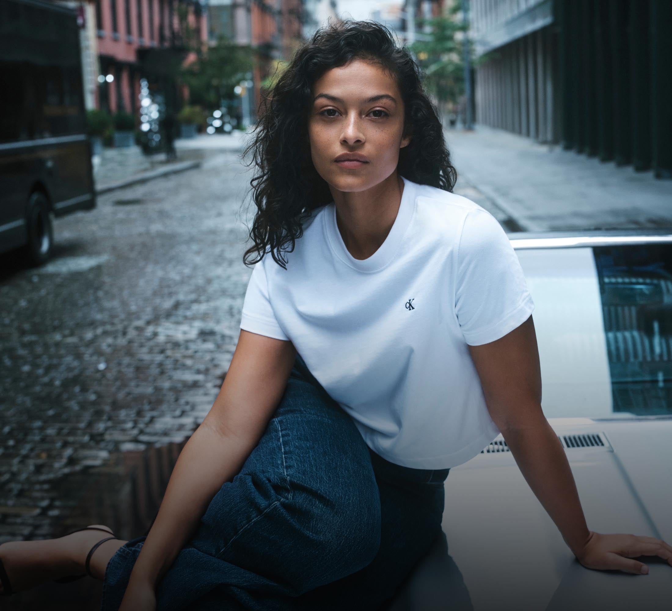 Woman sitting on the hood of a car wearing a white CK logo t-shirt and blue jeans