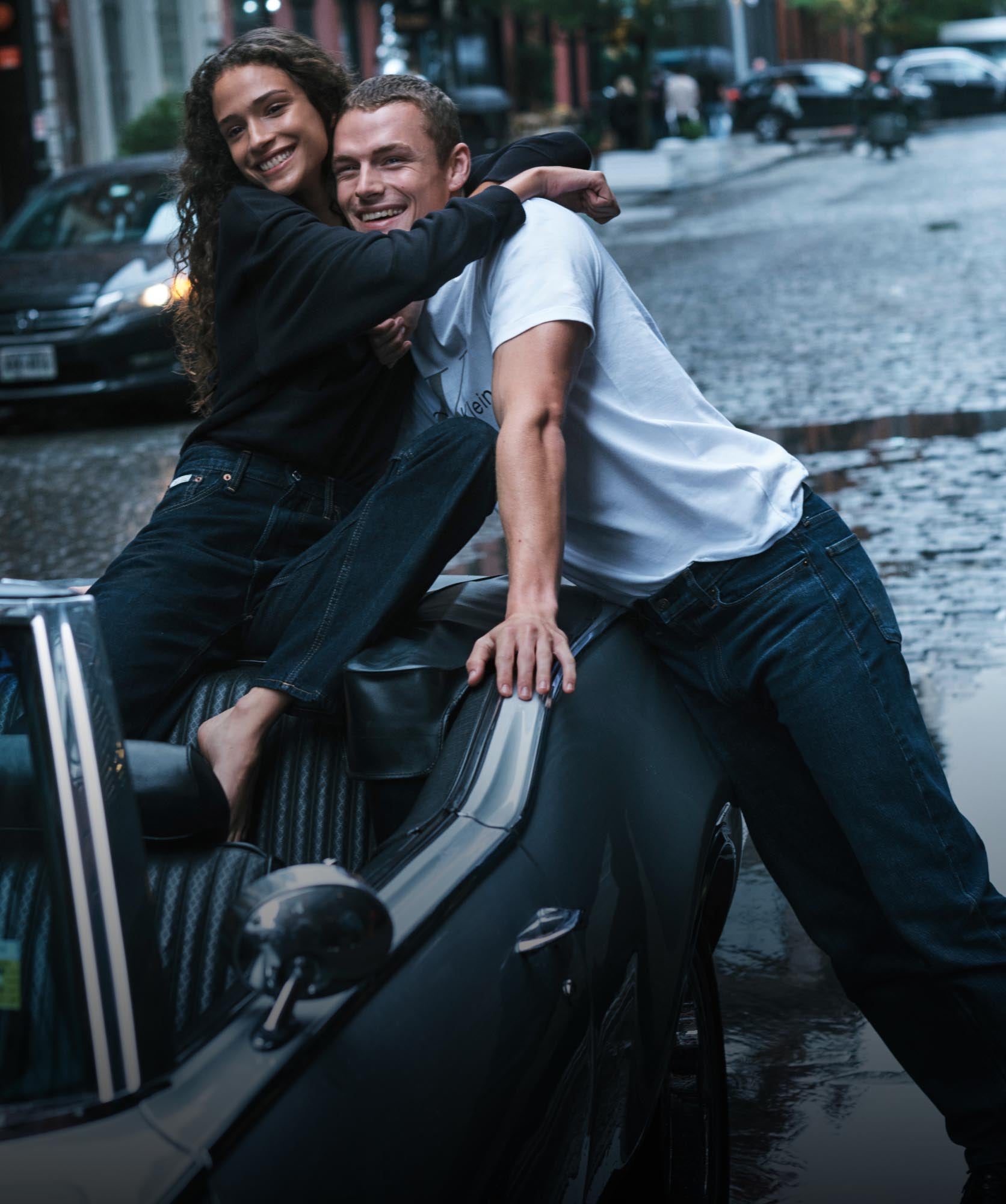 Woman wearing a matching sweat set sitting in a car wrapping her arms around a man wearing a t-shirt and jeans
