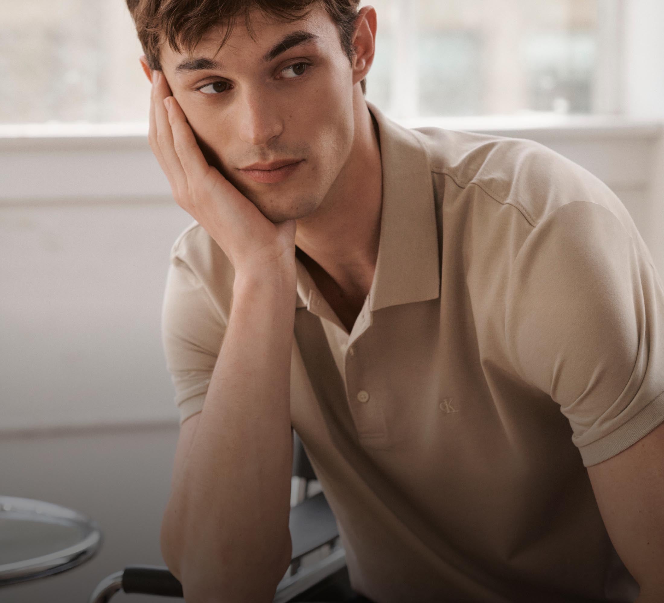 A male model posing with his hand resting on his chin wearing a tan Calvin Klein logo polo shirt