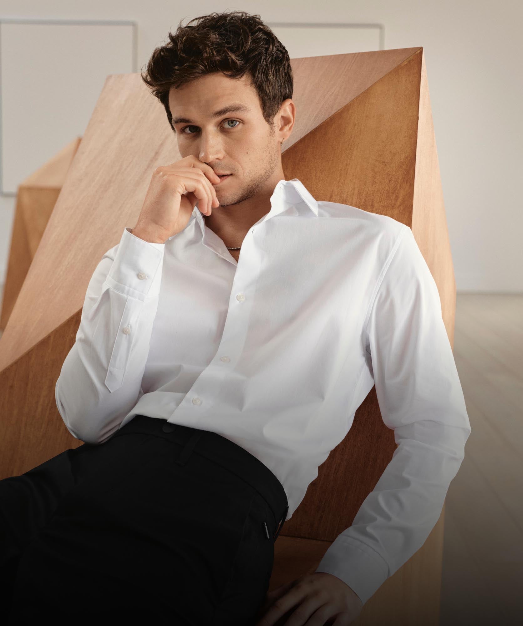 Brandon Flynn leaning back on a chair wearing a white button down shirt