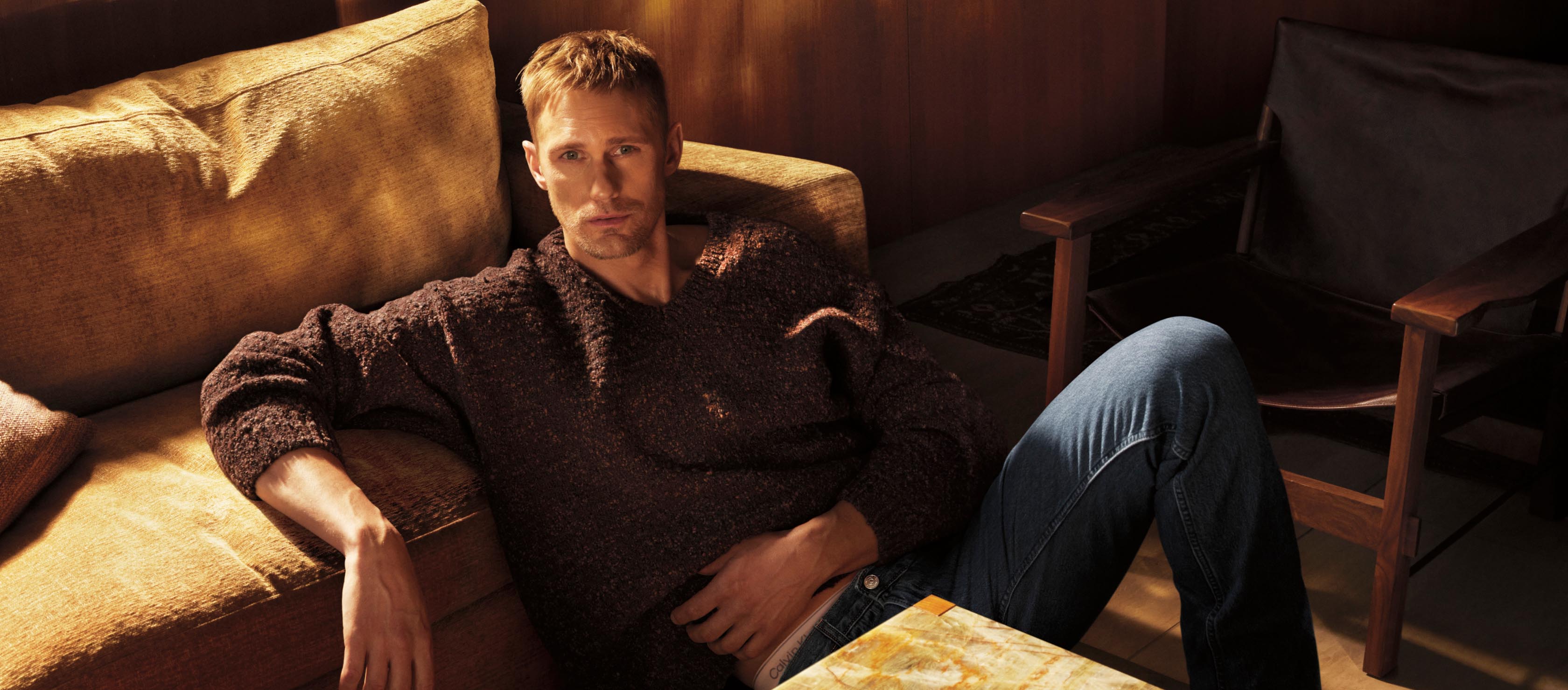 Alexander Skarsgard sitting on the floor leaning against a couch wearing a chunky brown sweater