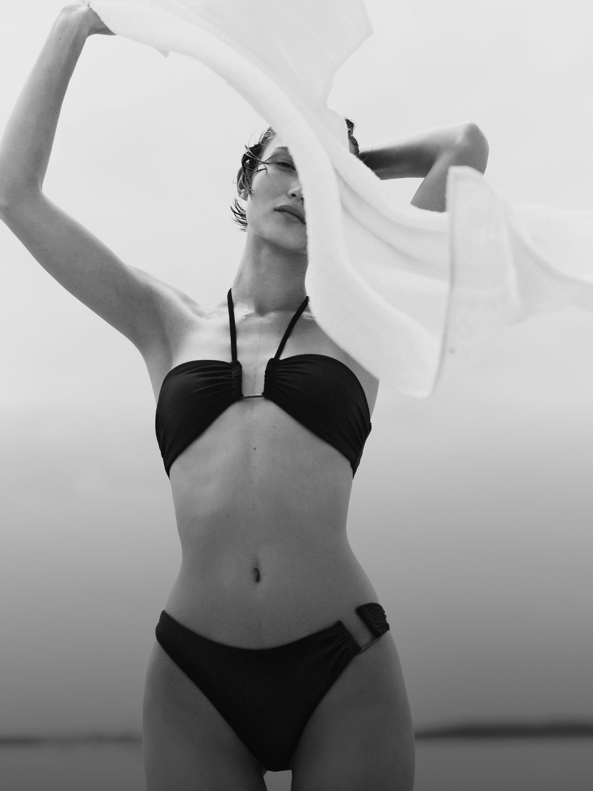 Black and white image of a model wearing a black bikini top and bottom at the beach.