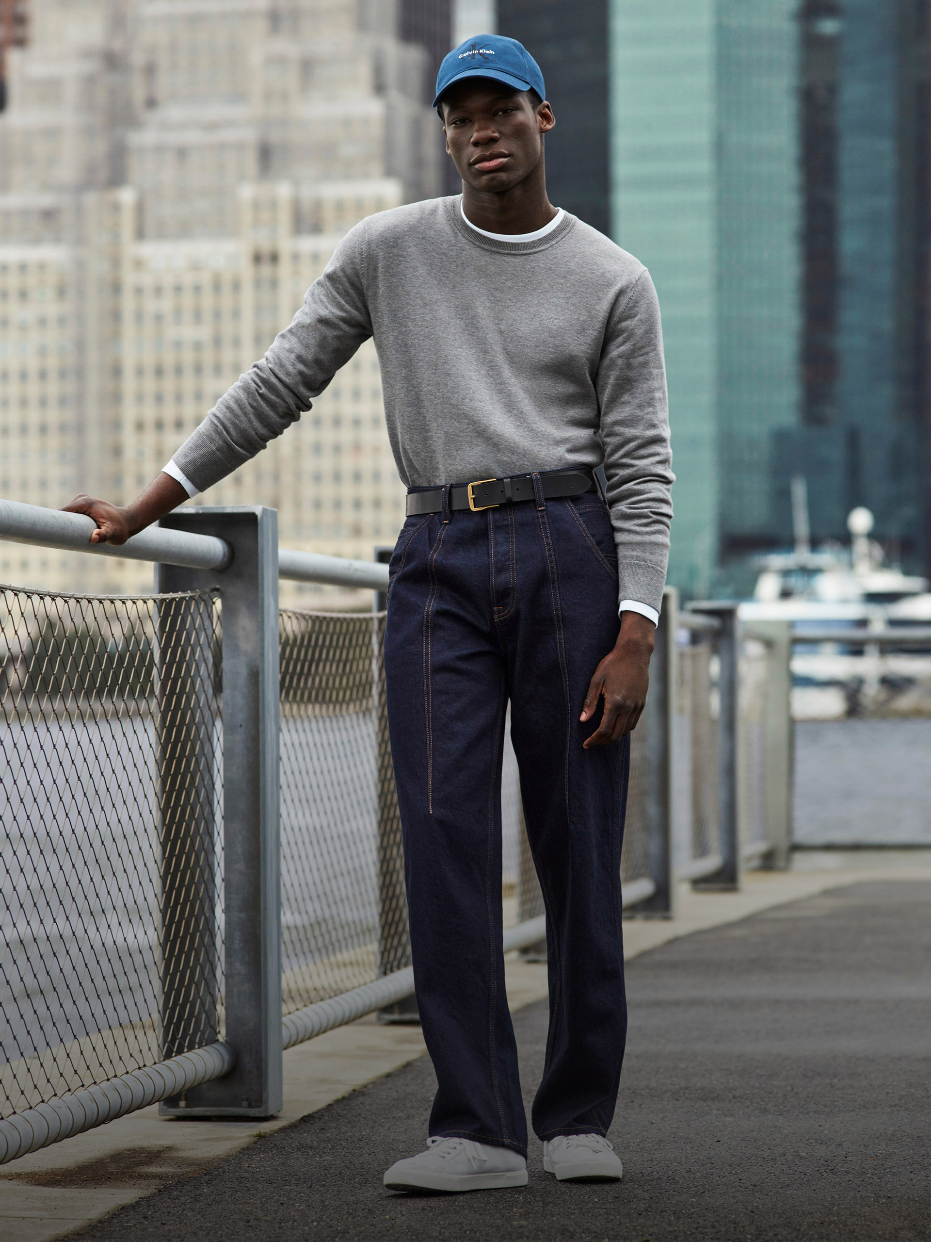 Model wearing a grey knit sweater with dark wash jeans while leaning on a fence in the park.