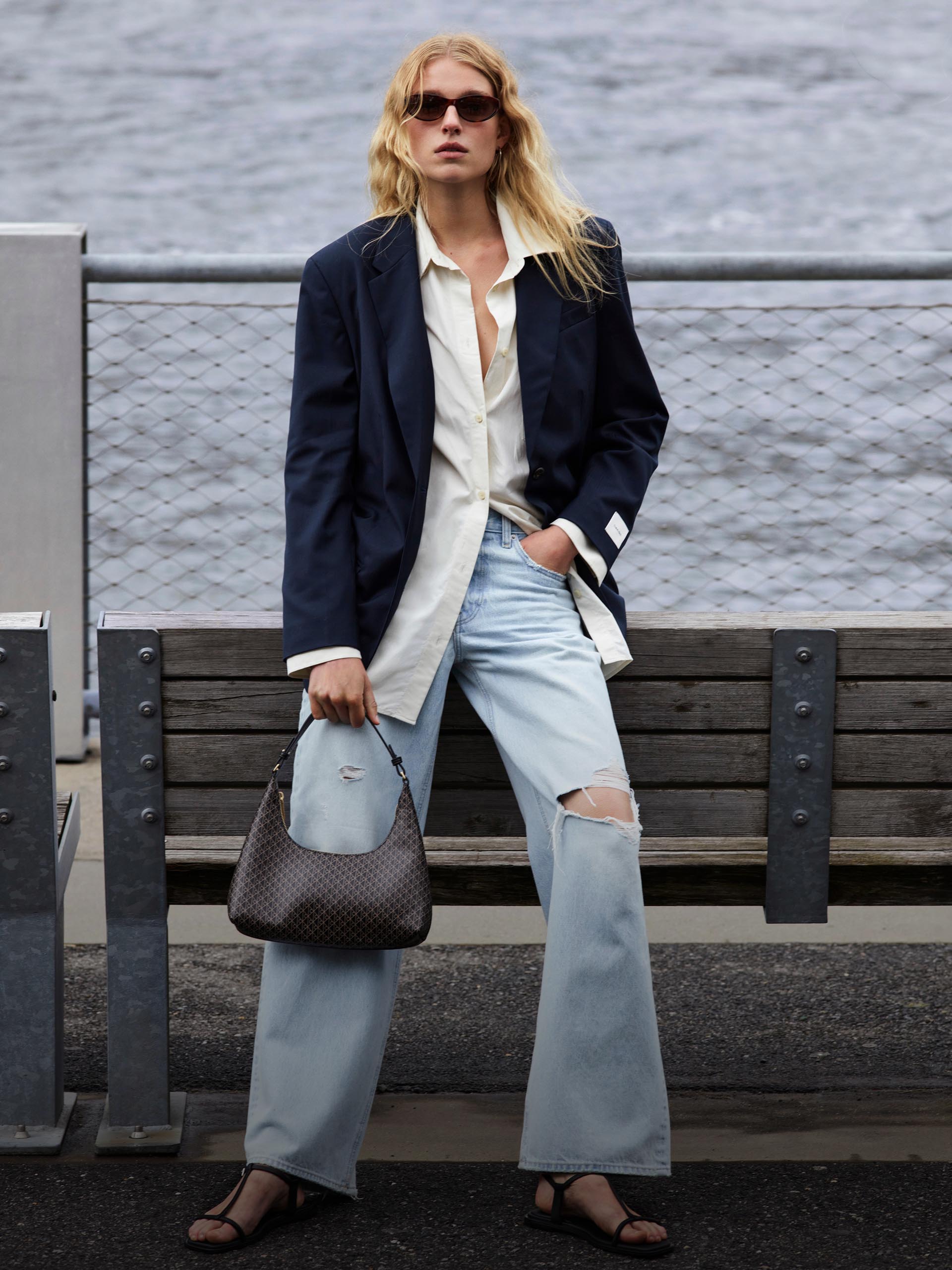 Model leaning on a bench wearing sunglasses, navy oversized blazer, white button-down shirt, and relaxed ripped jeans in a light wash.