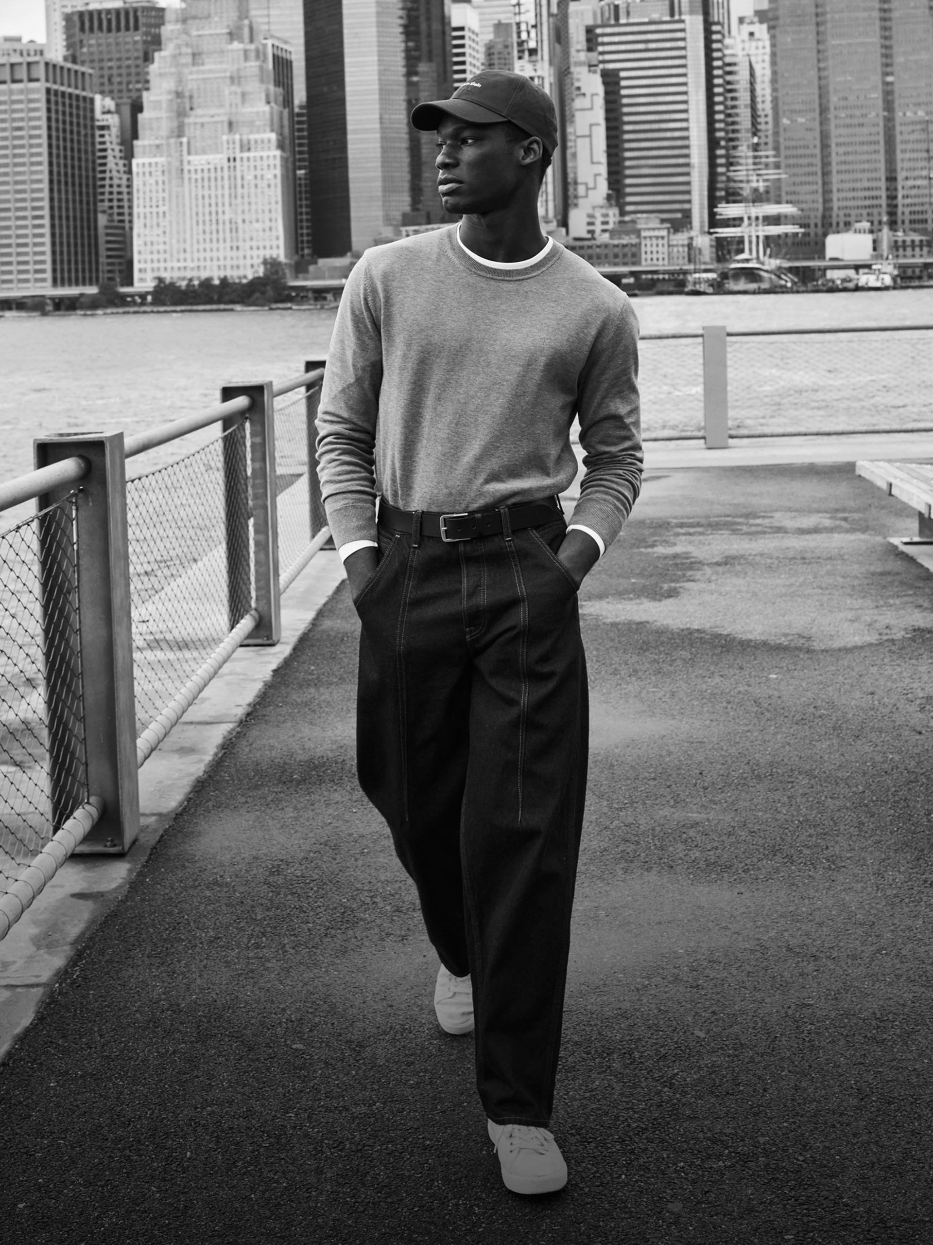 Model wearing a grey knit sweater with dark wash jeans walking in the park overlooking Manhattan.