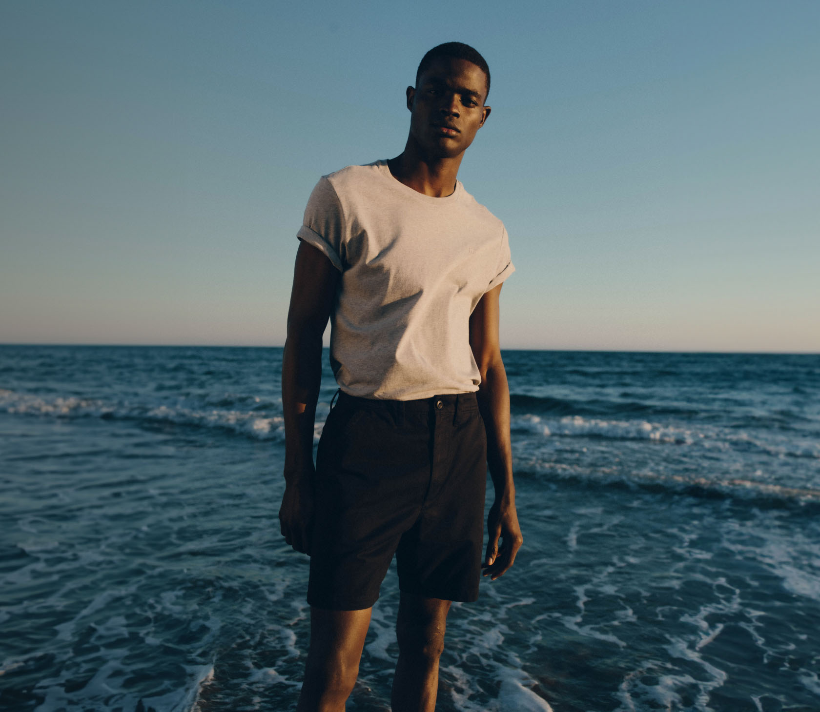 One image of a man standing in the ocean wearing a grey short sleeve t-shirt and blue chino shorts