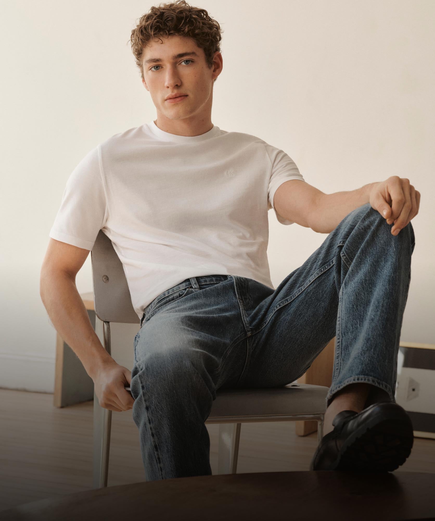 A male model leaning back in a chair with his leg up wearing a white t-shirt and medium wash blue jeans