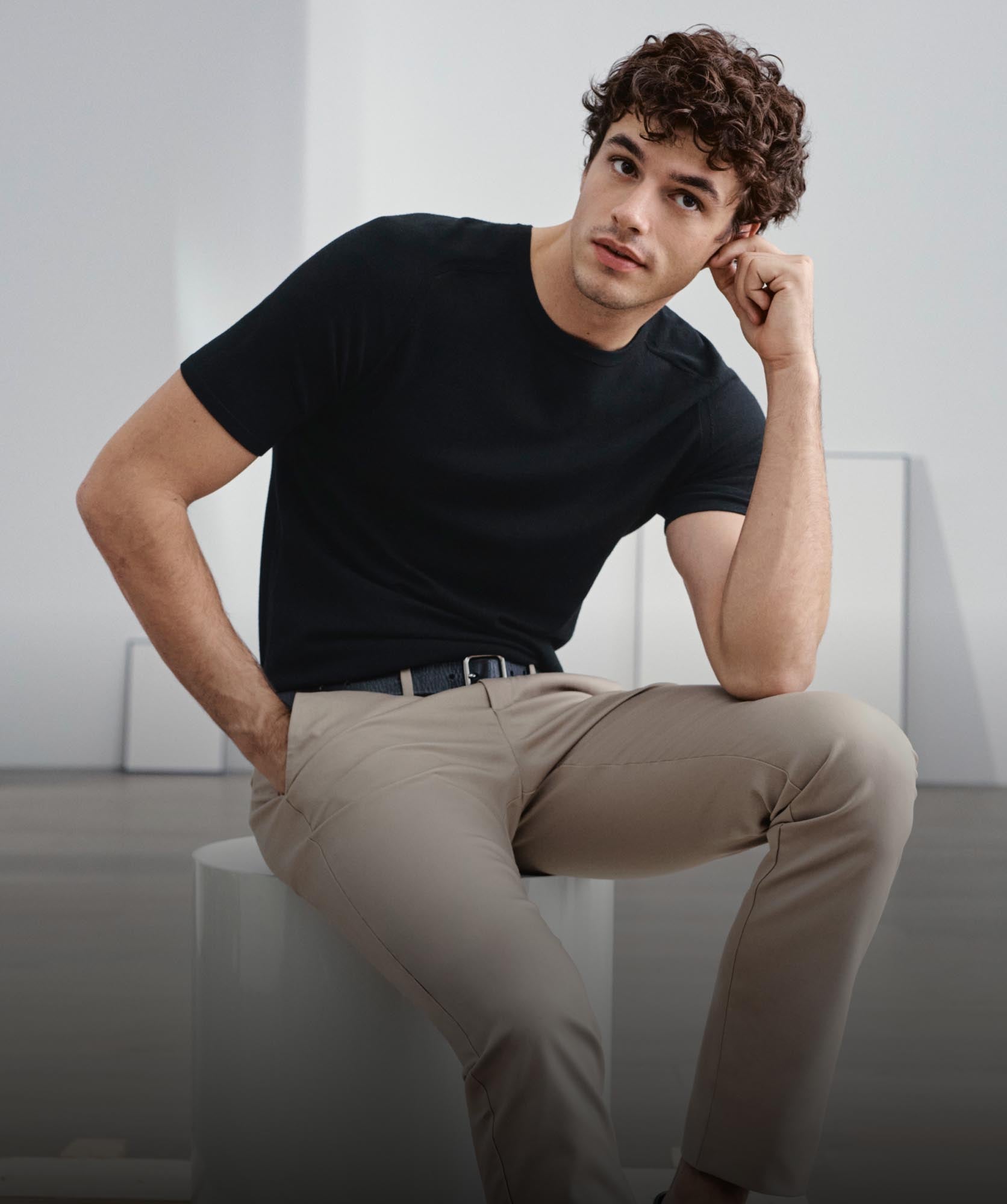 A model sitting down, posing with his elbow on his knee wearing a black t-shirt and khaki pants