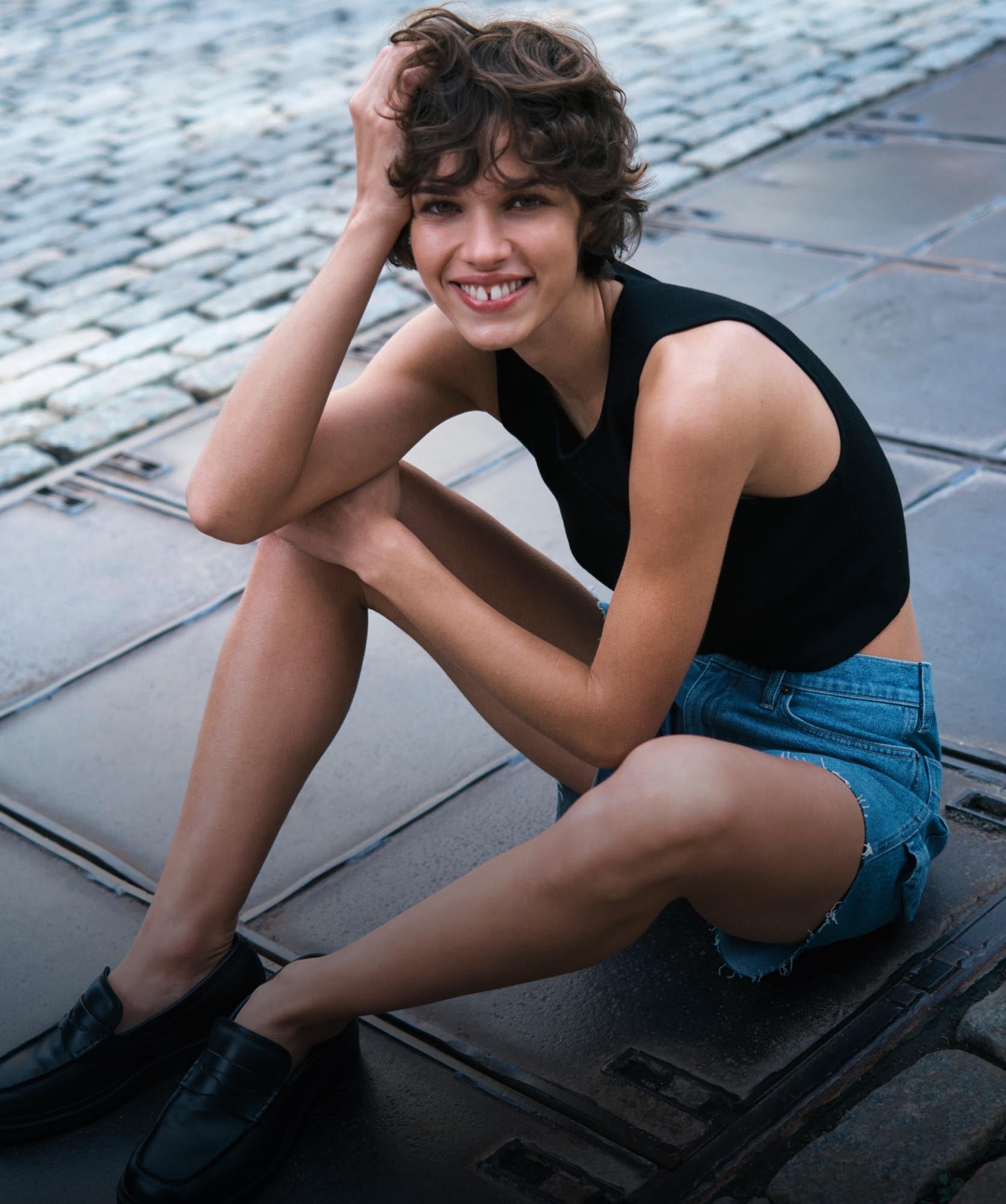 A model sitting on the ground posing with her hand in her hair wearing a black tank top and denim shorts 