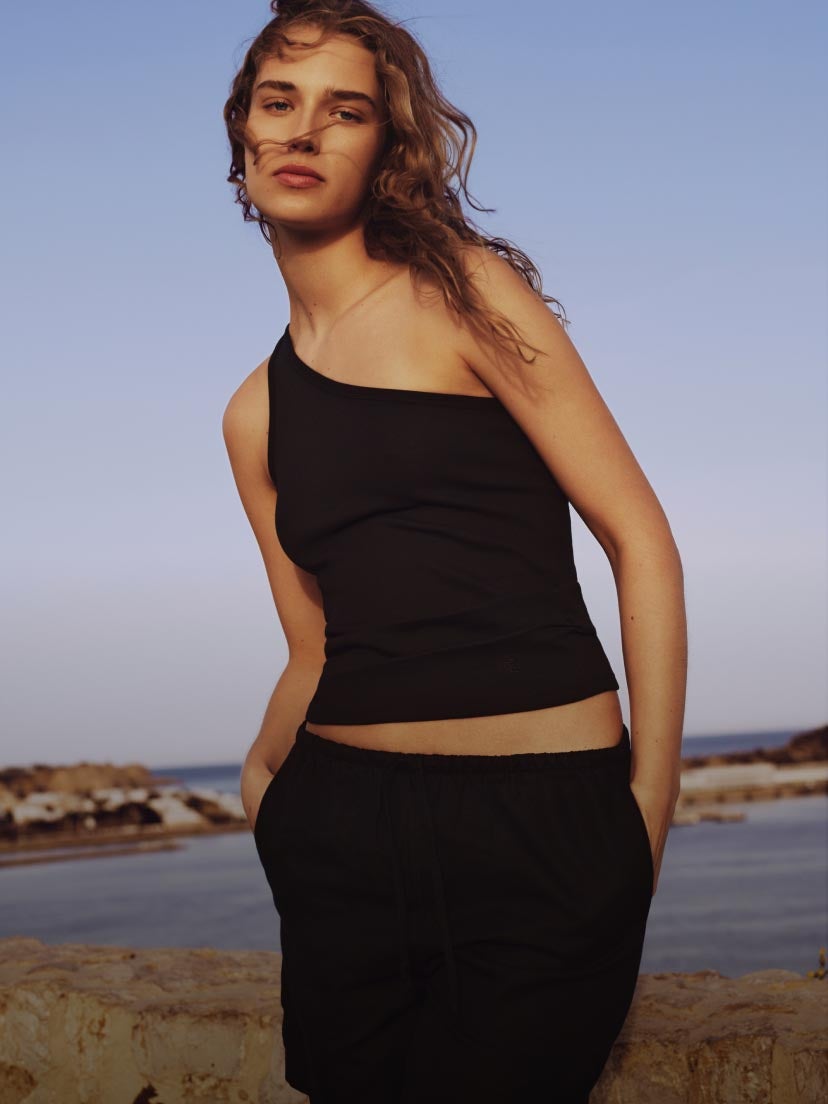 Model posing on a beach wearing a one shoulder black tank top