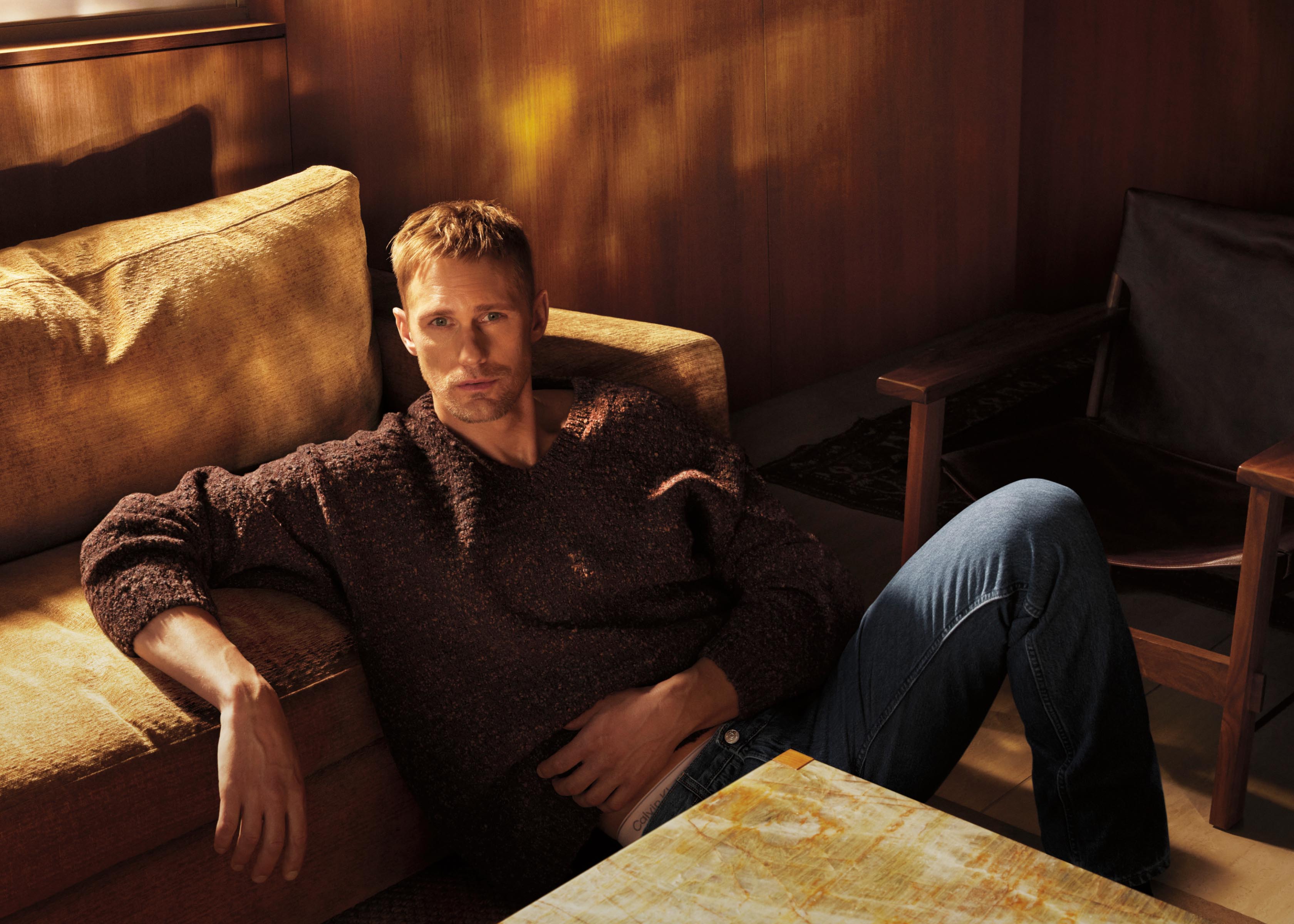 Alexander Skarsgard assis sur le sol adossé à un divan et vêtu d’un chandail brun épais