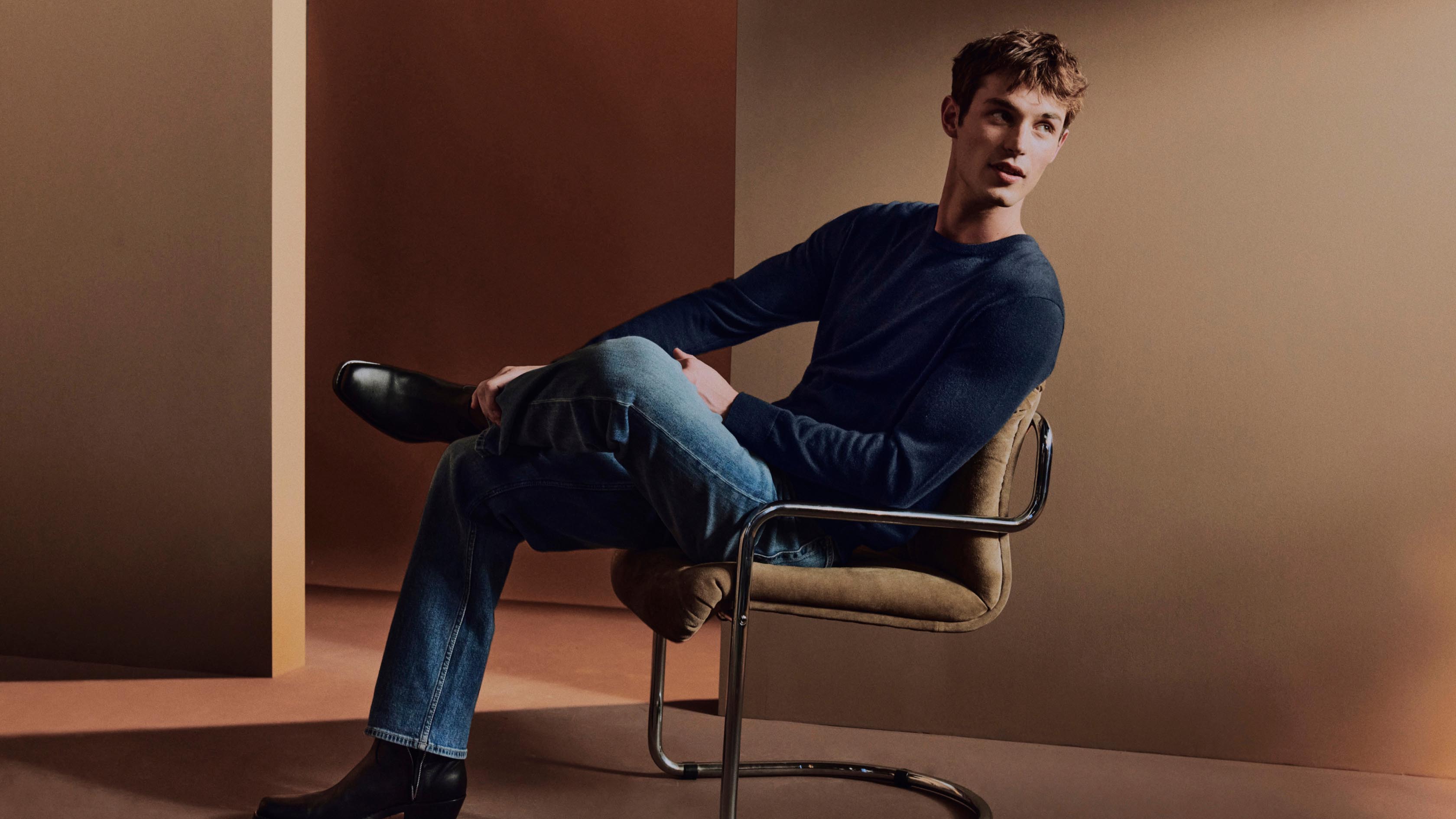 Model posing in a chair with his legs crossed wearing light blue denim jeans