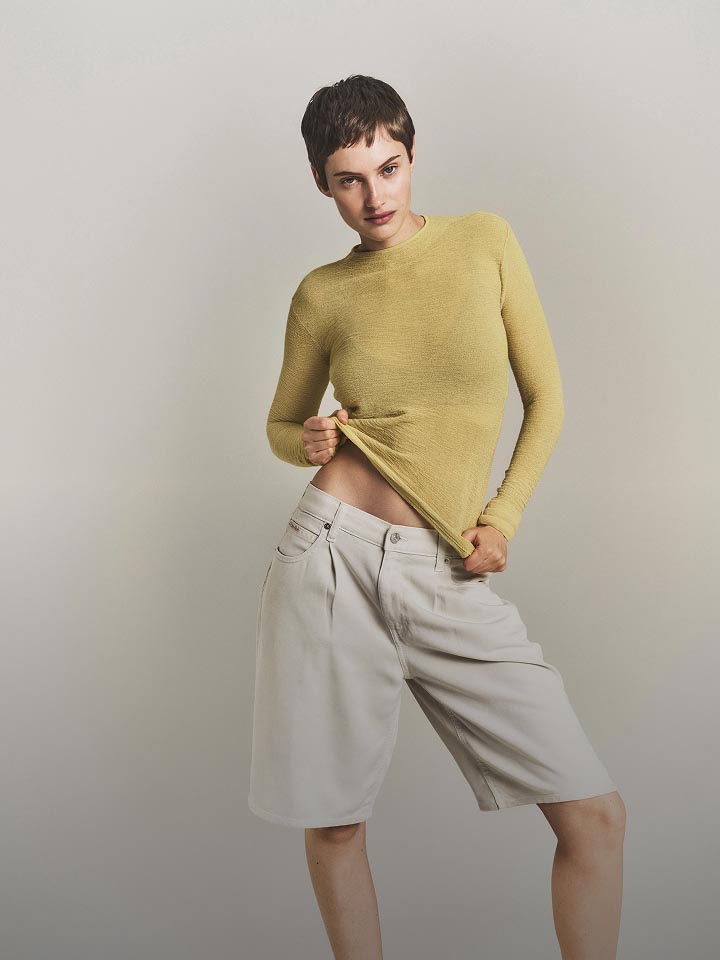 Model posing in a light yellow long sleeve sweater and white shorts.