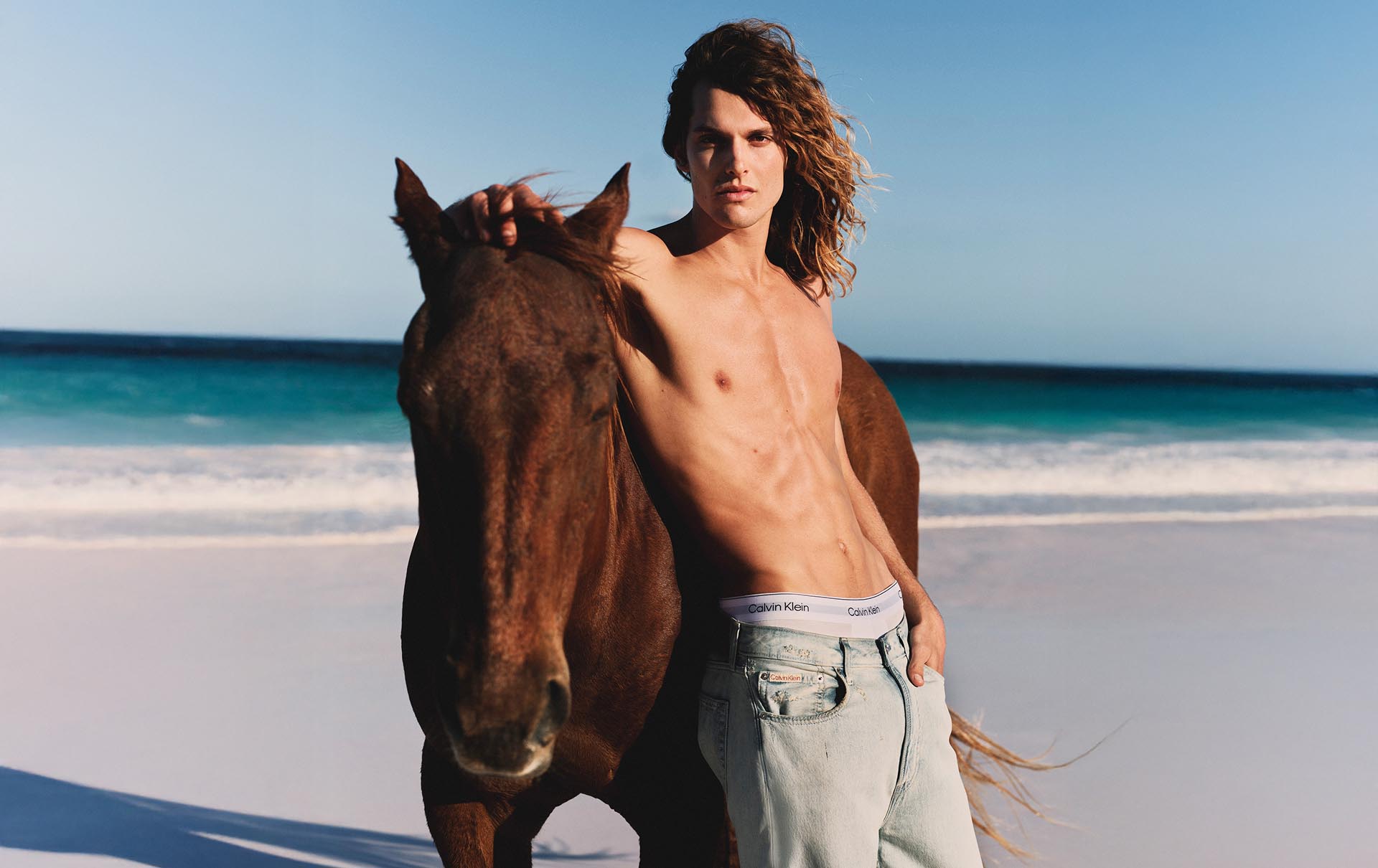 Model posing in a denim jeans on the beach with a horse.