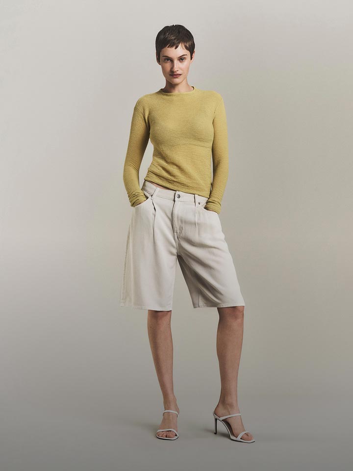 Model posing in a light yellow long sleeve sweater and white shorts.