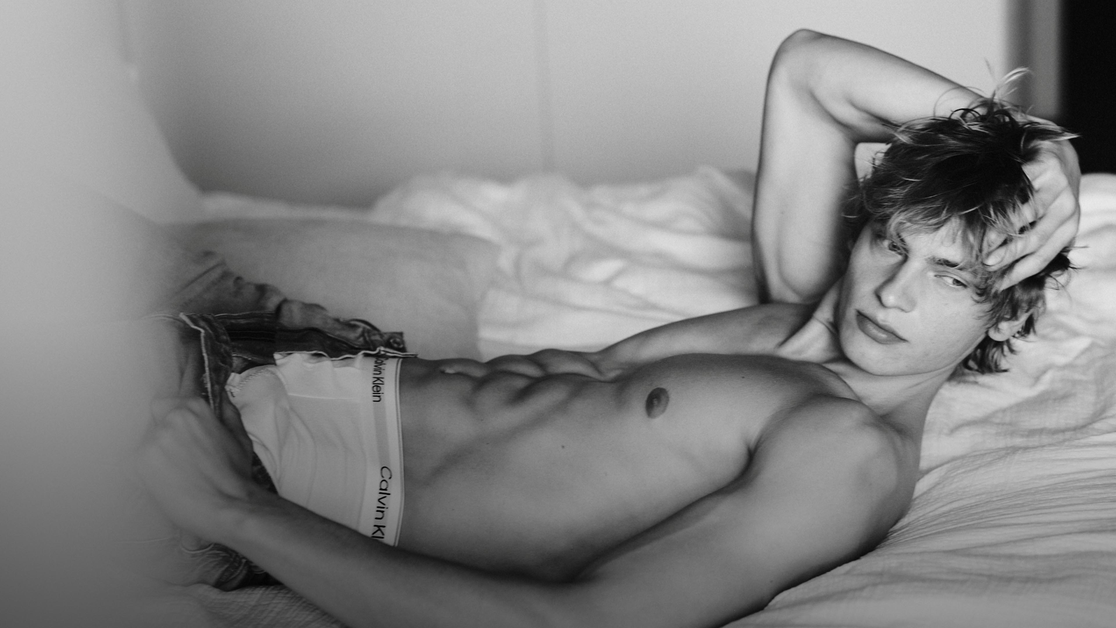 Model laying down on the bed in a black and white photo showing off their physique and the iconic Calvin Klein boxer brief under their blue jeans.