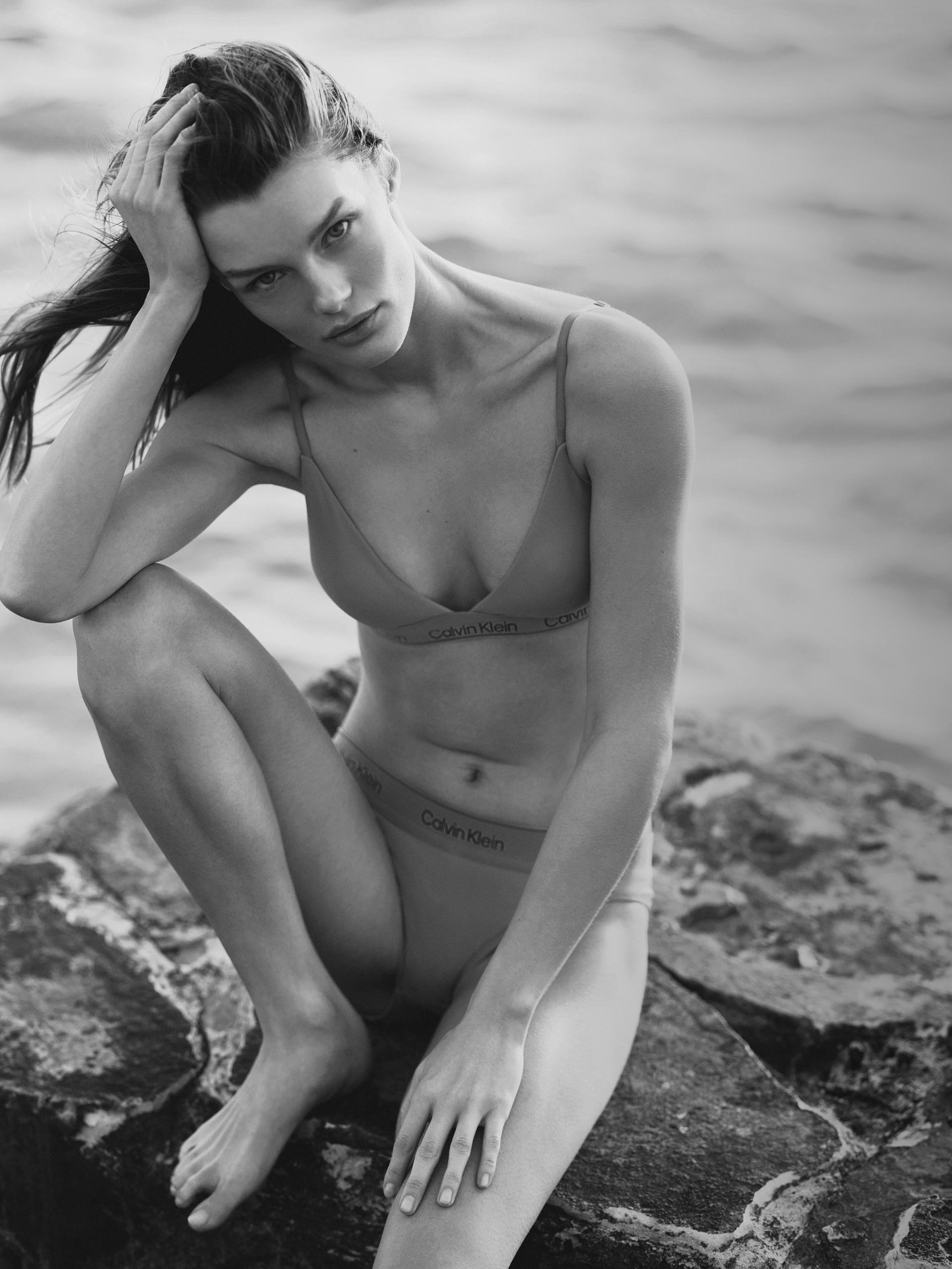 Black and white image of a model wearing a triangle bikini top and bottom with Calvin Klein logo across the bands. Model is sitting on a rock at the beach.