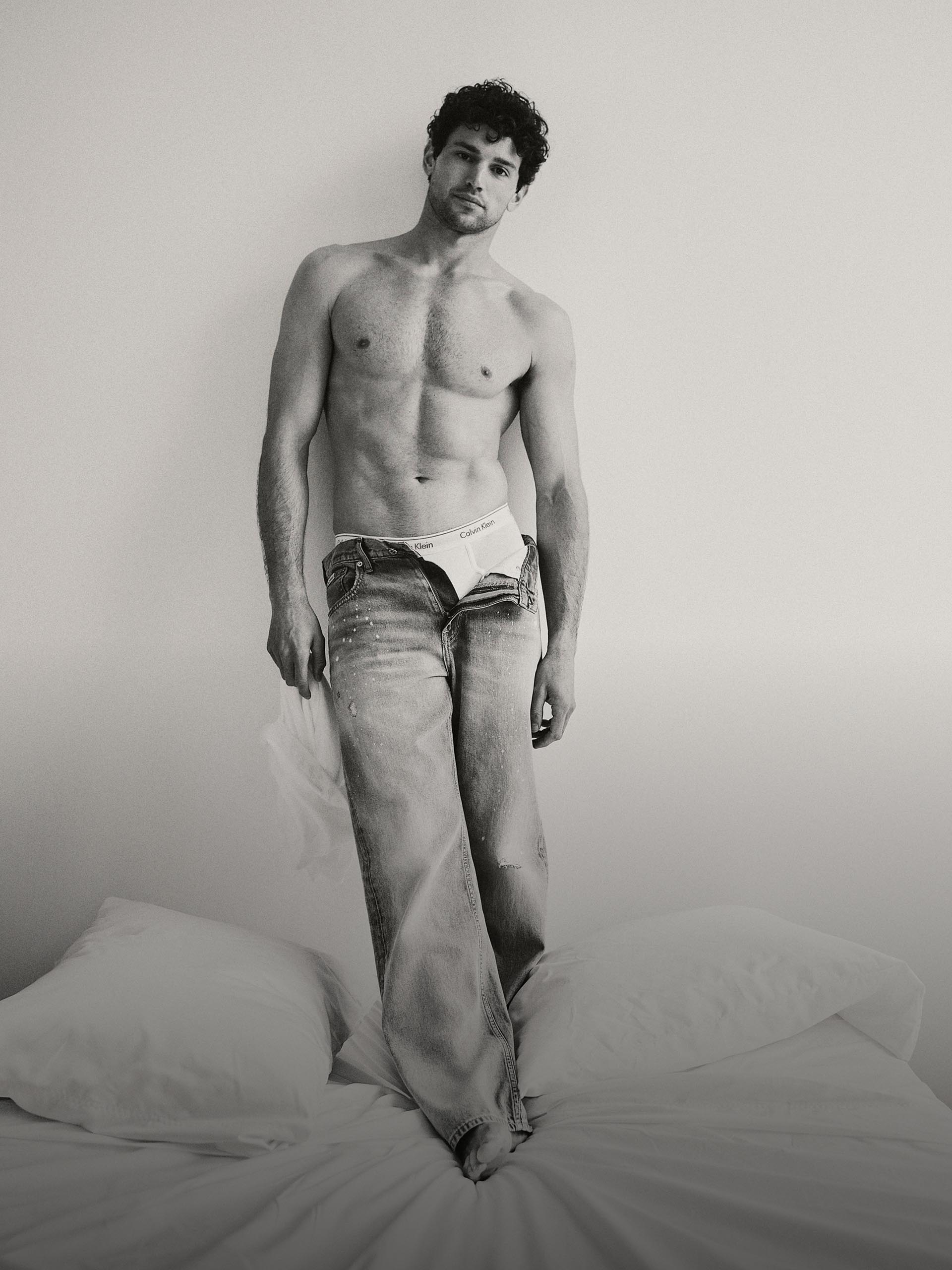 Black and white image of a model standing on a white linen bed wearing a half unzipped denim jeans showing off the iconic Calvin Klein white boxer brief.