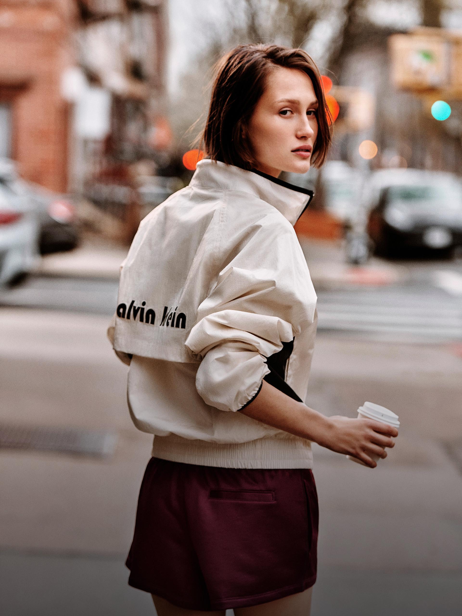 Model wearing a jacket and shorts while holding a coffee cup.