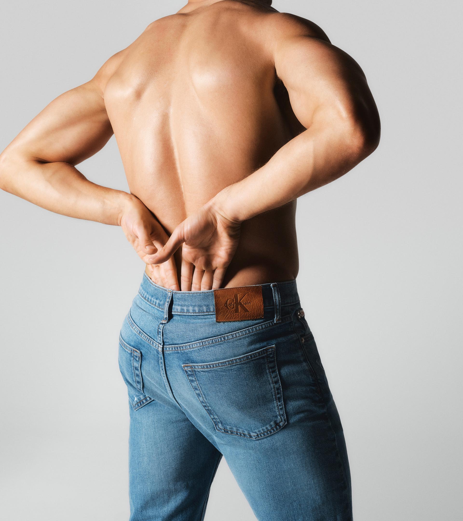 Model with his back to the camera only wearing a light wash jeans with the Calvin Klein logo patch.