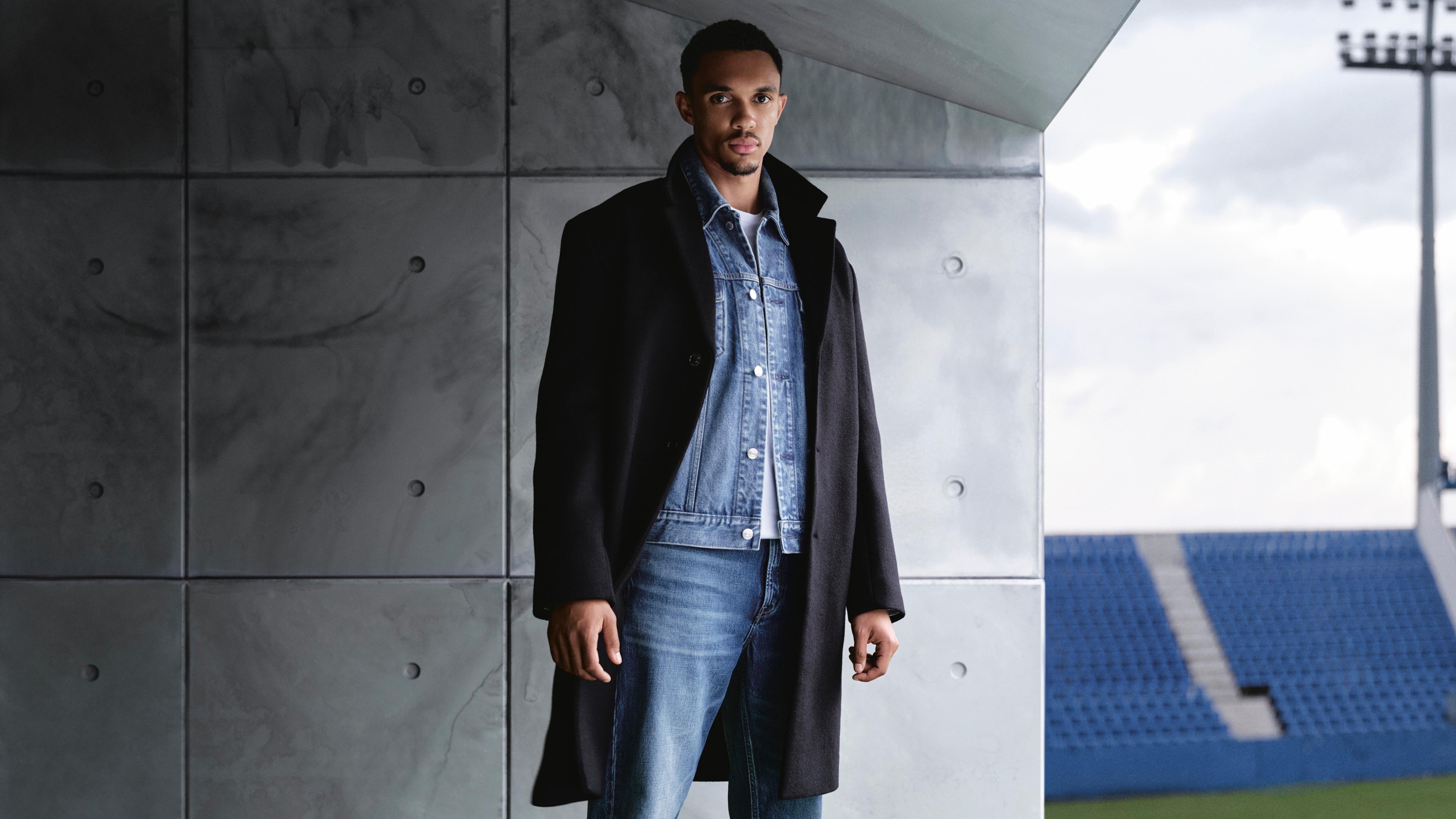 Trent Alexander-Arnold in a black wool coat over a denim jacket and jeans outfit in the football field.