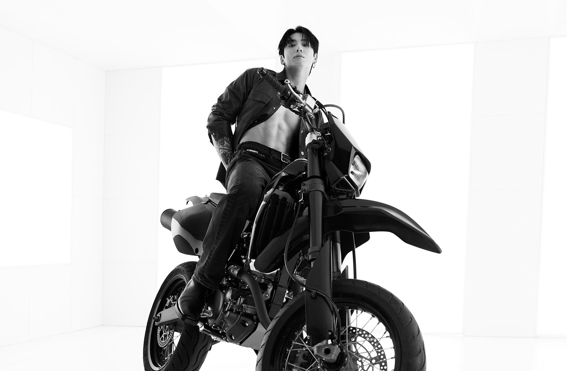 Jung Kook posing on a motorcycle in an unbuttoned leather shirt and jeans