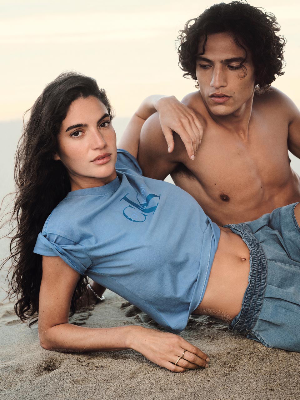 Model posing on a beach in a blue CK logo t-shirt