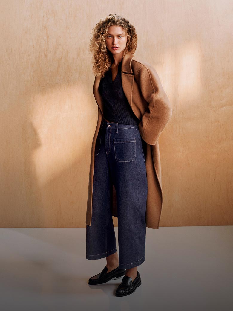 Model posing in a tan coat, black shirt, and high rise denim
